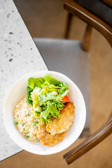 A white bowl with fried patties, fresh salad of lettuce and tomatoes, and creamy risotto with peas, placed on a speckled countertop near a wooden chair. A balanced, homemade meal with vibrant colors