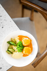 A bowl with mashed potatoes, sliced cucumbers, two pieces of food covered in orange sauce, and parsley garnish sits on a speckled countertop next to a wooden chair.