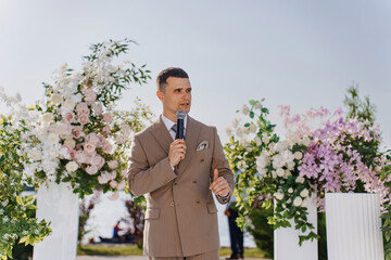 A man presenter in a beige suit leads a wedding ceremony in the park. 