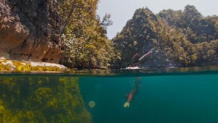Freediver swims underwater in the closed lake. Splitted above and underwater view of the diver swimming in the lake surrounded by limestone mountains. West Papua, Misool, Indonesia