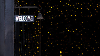 A, WELCOME, sign hangs on the dark black pole with a small bell. The background sparkles with numerous soft yellow lights, creating a warm and inviting atmosphere.