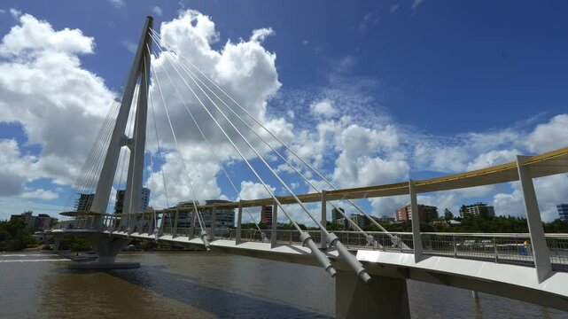 New Kangaroo Point Green Bridge over the Brisbane River, time-lapse shot capturing pedestrians crossing and ferries cruising on the waters on a sunny day under a blue sky with fluffy cumulus clouds.