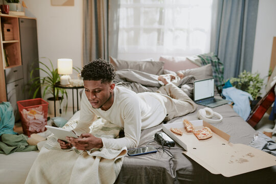 Young man relaxing on bed while using tablet in a cluttered room filled with personal items and various devices, creating a casual, relatable indoor scene