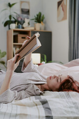 Woman enjoying peaceful moment while reading book on bed, surrounded by cozy and warm decor in...