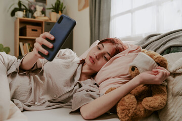 Young woman relaxing on cozy bed, engaging with her smartphone. She seems engrossed in her activity...