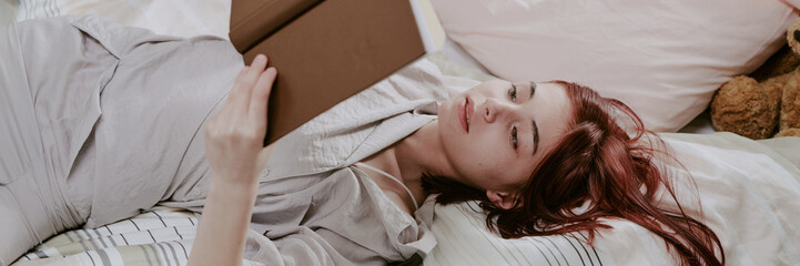 Woman with red hair immersing herself in a book while lying on a bed surrounded by white linens and...