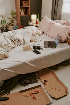 Fototapeta Disorganized bedroom with unmade bed, scattered pillows, open laptop, and pizza boxes. Shelves and dresser filled with books and decor items in background