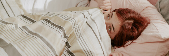 Fototapeta premium Young child peering from beneath striped bedding, revealing partial face while laying in bed. Child's hair is reddish-brown, with bedding environment cozy and comfortable
