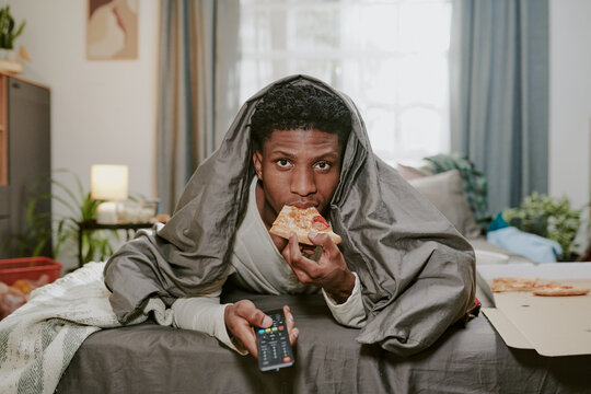 Young man eating pizza while watching TV under cozy blanket in living room filled with various household items and soft lighting