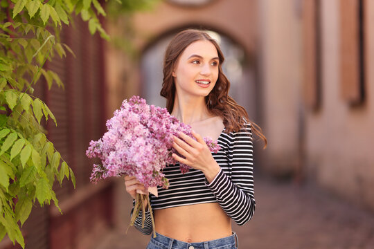 Fototapeta Smiling woman with bouquet of lilac flowers near tree on city street