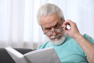 Senior man with vision problem reading book on sofa at home