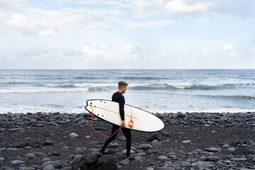 Surfer walking on black volcanic beach with surfboard under his arm