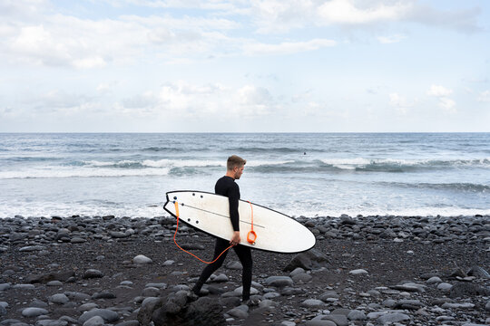 Surfer walking on black volcanic beach with surfboard under his arm