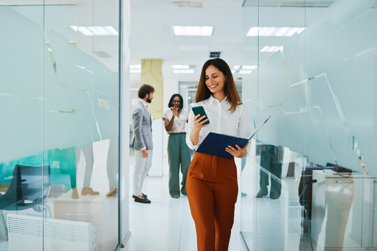 Smiling businesswoman walking through office corridor using smartphone and holding documents