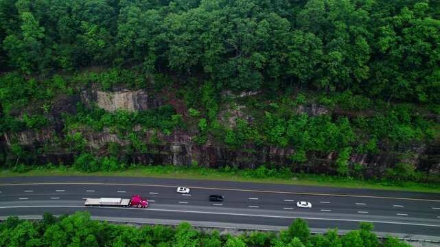 Aerial drone tracking shot of a red flatbed semi truck hauling cargo along I-24 through Monteagle&rsquo;s forested cliffs, highlighting logistics.