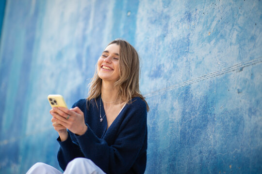 Joyful young woman sitting against a blue wall, laughing while using her phone - Powered by Adobe