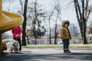 Obraz premium A child wearing a winter coat stands near a yellow slide in a park, with a dog and another individual nearby amidst the outdoor scenery.