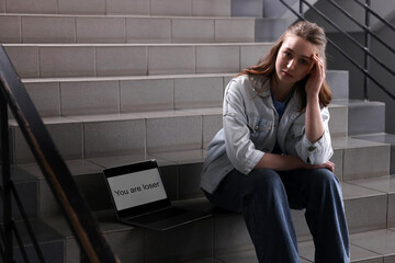 Cyber bullying. Upset teenage girl and laptop with text You Loser on stairs indoors