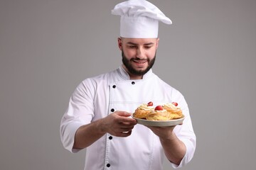 Happy confectioner in uniform holding delicious profiteroles with strawberries on light grey background