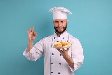 Happy confectioner in uniform holding delicious profiteroles with strawberries and showing ok gesture on light blue background