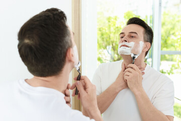 Handsome man shaving with razor near mirror at home