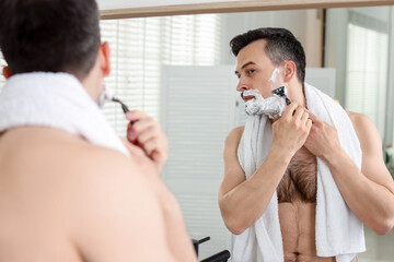 Handsome man shaving with razor near mirror at home