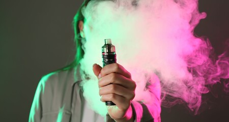 Woman using electronic smoking device in neon lights on grey background, selective focus