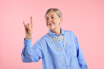 Senior woman wearing elegant pearl jewelry and making rock gesture on pink background