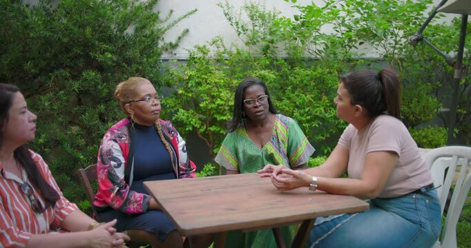Four diverse women gather in backyard around wooden table, actively engaged in meaningful outdoor conversation with expressive body language