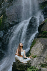 Obraz premium young woman meditating peacefully near a waterfall surrounded by rocks and lush greenery in natural outdoor setting during daytime