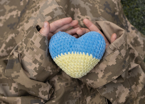 children's hands in a large camouflage uniform hold a knitted yellow and blue heart. Support Ukraine with charity. Defender of Ukraine Day. Independence Day. Faith in the Armed Forces of Ukraine