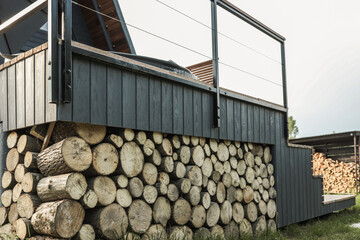 Stacked firewood stored under a dark wooden terrace with black railing, showcasing sustainable...
