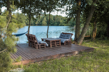 Wooden deck with handmade pallet furniture and soft cushions by a calm lake, surrounded by trees. A...