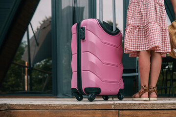 Close-up of young girl in checkered dress standing with pink suitcase and straw hat, ready for...