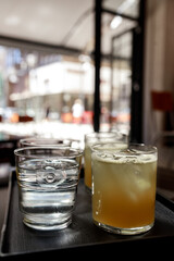 Beverage glasses placed on black tray inside cafe with bright outdoor city scene visible in background, focus on foreground drinks.Glasses of iced juice and water on tray with blurred city street view
