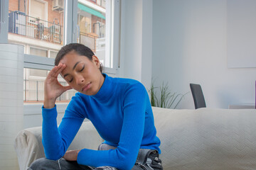 Latin young woman touching her head with headpain, stress sitting on the couch at the livingroom ...