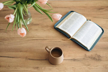 Open Bible on a wooden table and ceramic coffee mug and a vase with tulips.