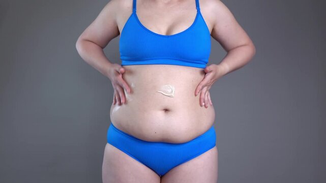 Abdominal massage, tummy tuck and flabby skin on a fat belly, woman doing self-massage with cream on gray background, plastic surgery concept