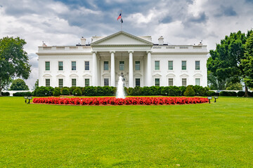 White House under a cloudy sky. Large mowed lawn with red flowerbed and fountain.