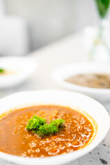 A close-up of bowl of tomato soup garnished with fresh parsley, placed on a light-colored surface. The background includes blurred elements of other dishes, creating a cozy and appetizing atmosphere.