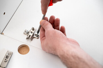 Hand using a screwdriver to fasten a metal hinge bracket on a white cabinet panel