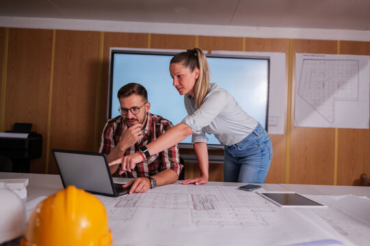 Two architects are collaborating on a project, examining blueprints and using a laptop in their office, showcasing teamwork and expertise in architectural design