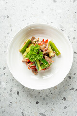A fresh and colorful salad featuring sliced cucumbers, red bell peppers, and tender pieces of meat, garnished with parsley, served in a white bowl on a speckled countertop.