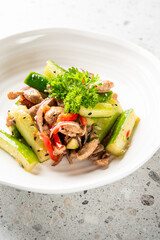 A fresh and colorful salad featuring sliced cucumbers, red bell peppers, and tender pieces of meat, garnished with parsley, served in a white bowl on a speckled countertop.