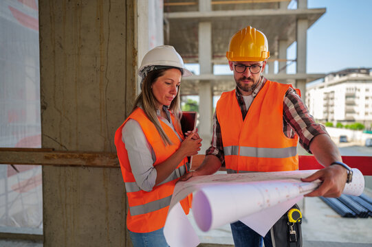 Two construction workers, a man and a woman in safety vests and helmets, reviewing a blueprint on a construction site while discussing project plans