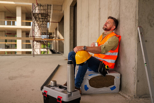 Construction worker relaxing on insulation material, enjoying a hot drink from a thermos, with a hard hat resting on his leg, showcasing dedication and hard work