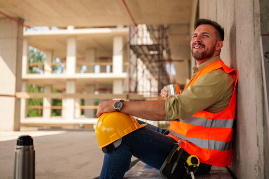 Construction worker wearing safety vest and hard hat enjoying a coffee break, sitting and resting against a concrete wall at the construction site, taking a moment to relax and recharge