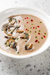 Creamy mushroom soup in a white bowl, garnished with sautéed mushrooms, fresh herbs, and drops of red and green sauces, set on a speckled gray countertop. Close-up, top-down view, artistic