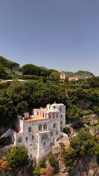 Drone view of Villa Cimbrone at golden sunset, perched dramatically on a cliff over Salerno Bay. Breathtaking Amalfi Coast scenery, sheer drop, historic architecture, destination wedding in Italy