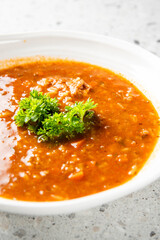 A bowl of rich tomato soup garnished with fresh parsley on a speckled countertop. The soup is hearty, with visible chunks of vegetables and meat, offering a warm and comforting meal.